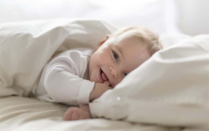 happy smiling 5 month old baby with finger in his mouth lying under a duvet cover on bed