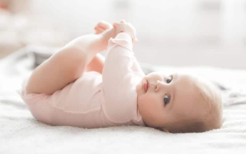 precious 5 month old baby in pink onesie lying on flat bed holding hands looking with blue eyes at the camera