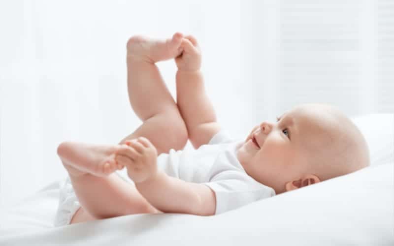 Happy 5 month old baby lying on bed with white sheets holding his feet and smiling